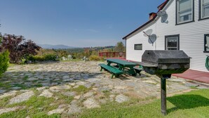 A stone patio with picnic tables and a barbecue grill in front of a white building with a red roof. - 'John's Farmhouse in Mount Snow' on 120 Acres! (Whitingham)