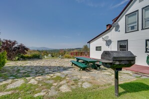 Een terras van natuursteen met picknicktafels en een barbecue voor een wit gebouw met een rood dak.