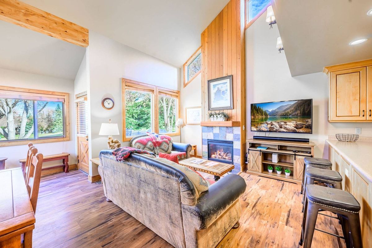 A cozy living room with a fireplace, wooden furniture, and large windows offering a view of trees.