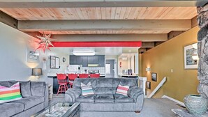 Ein Wohnzimmer mit grauem Sofa, Couchtisch und rotem Teppich. Im Hintergrund befindet sich eine Küche mit roten Stühlen und einer weißen Treppe.