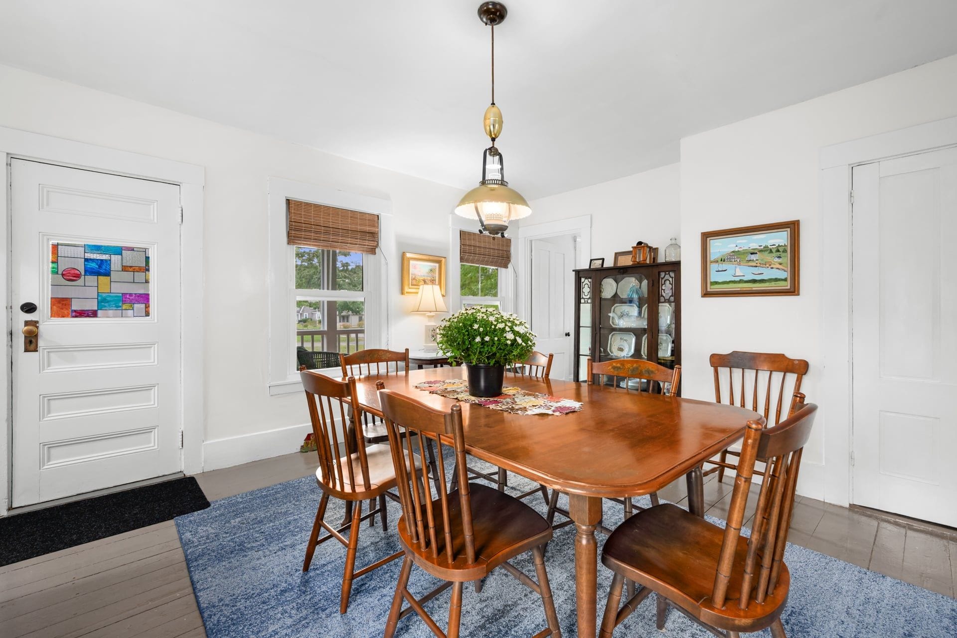 A dining room with a wooden table, chairs, a hanging light fixture, a potted plant, and a window with blinds.