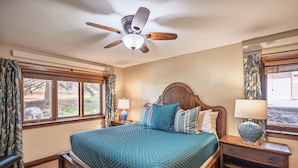 Un dormitorio con cama, cabecera de madera, dos mesitas de noche con lámparas, ventilador de techo y ventanas grandes con cortinas.
