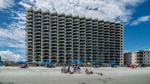 Un hotel di grandi dimensioni con numerosi balconi, situato vicino a una spiaggia con ombrelloni e lettini.
