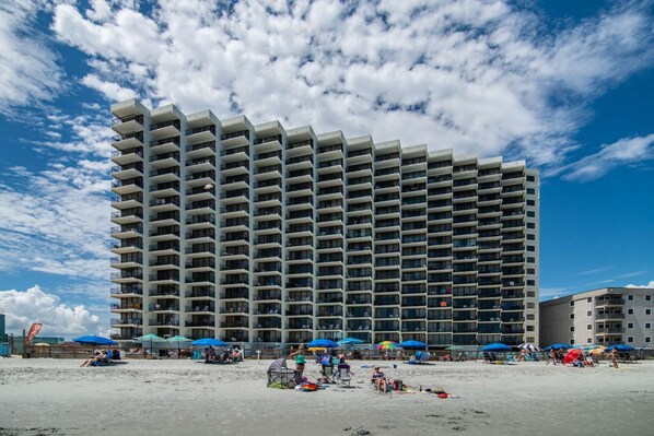 A high-rise hotel with multiple balconies, located near a beach with umbrellas and beach chairs.
