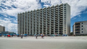 Un hotel moderno con molti balconi, situato vicino a una spiaggia con ombrelloni e persone.