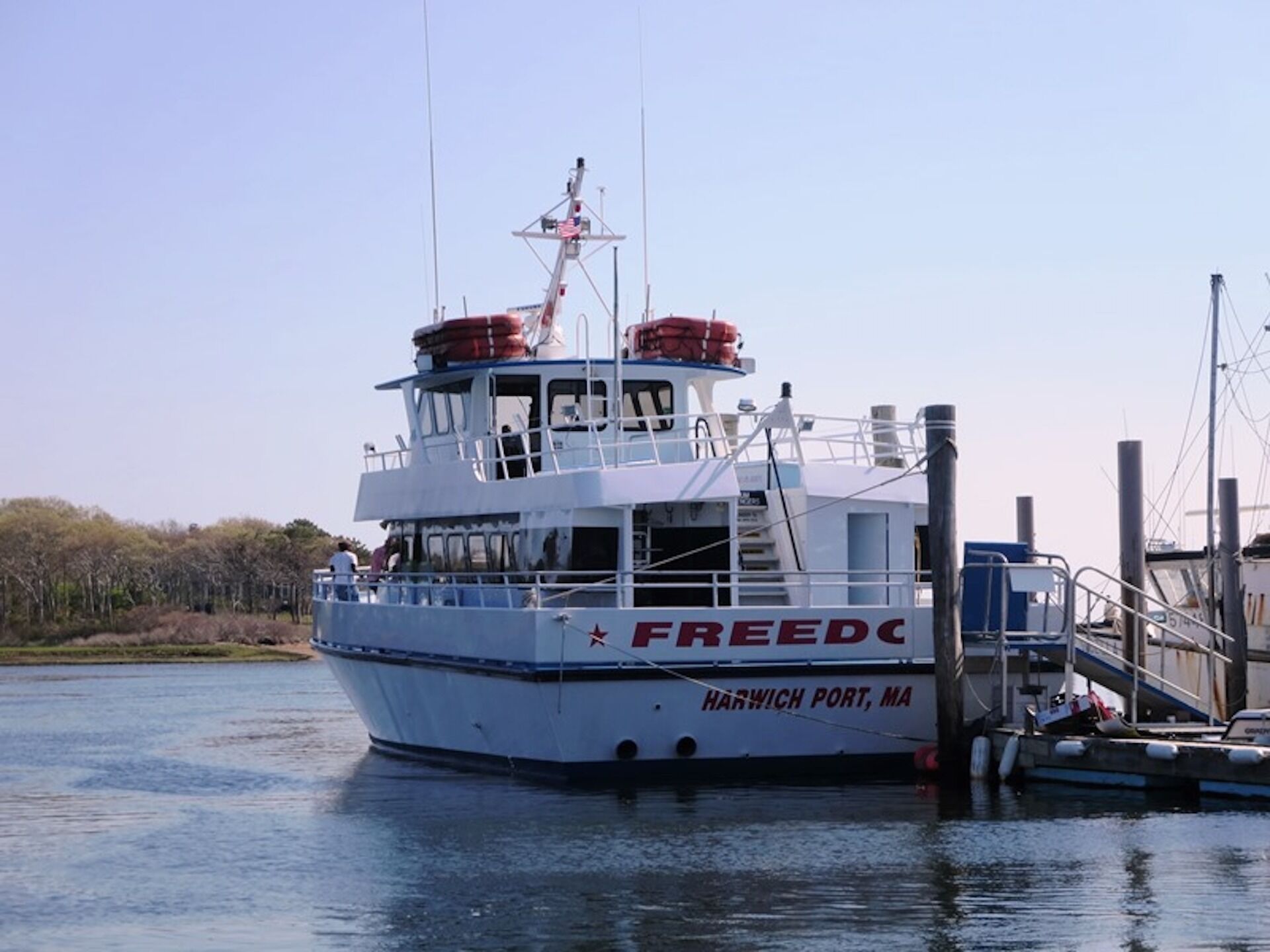 A white boat with 'FREEDOM' and 'HARWICH PORT, MA' written on it, docked at a pier with a clear sky in the background.