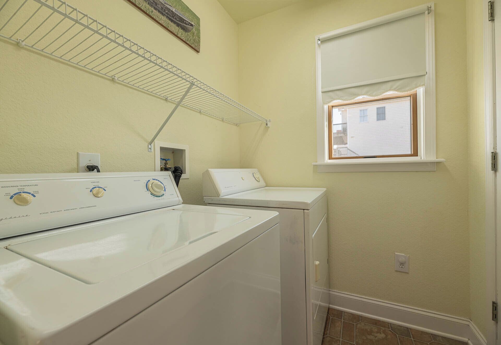 A laundry room with two white washers and dryers, a metal drying rack, a window with blinds, and a small electrical outlet on the wall.
