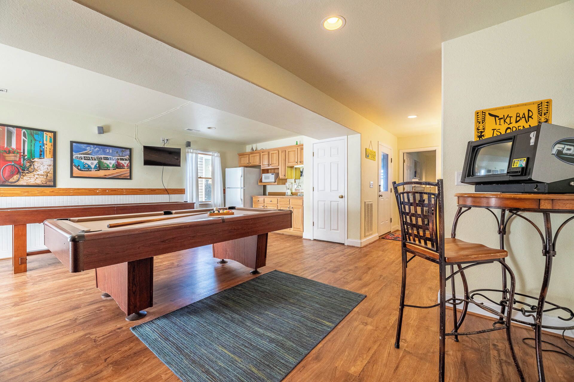 A spacious living area with a pool table, a bar area with a TV, wooden cabinets, and a kitchen in the background.