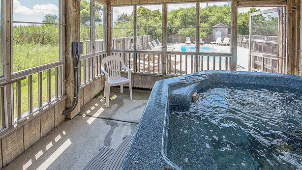 Een hottub in een houten chalet met grote ramen en uitzicht op een zwembad en bomen.