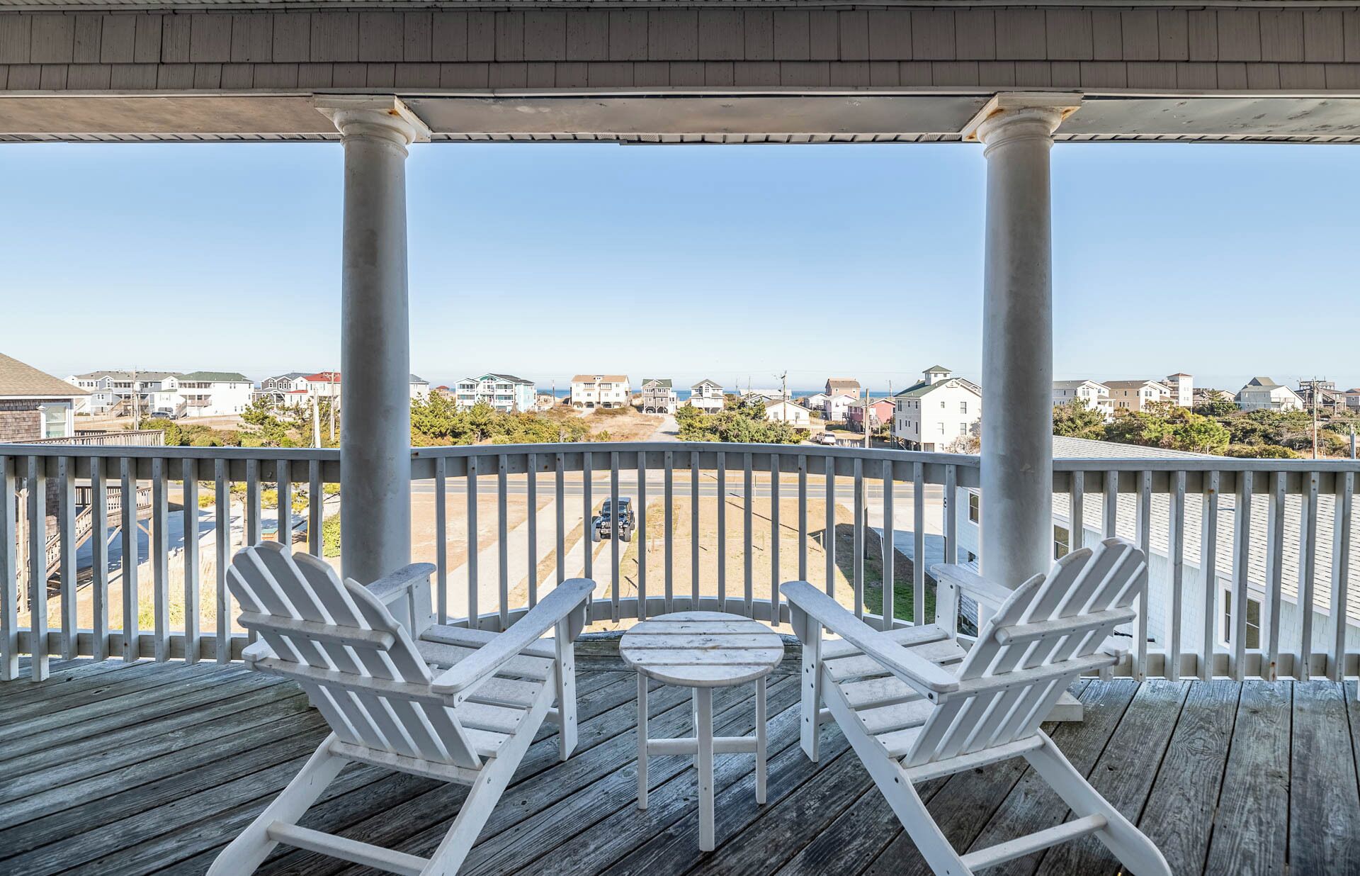 Um deck de madeira com duas cadeiras Adirondack e uma pequena mesa, com vista para uma área residencial com casas e um céu claro.