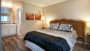 A bedroom with a large bed, a bedside table with a lamp, a wall-mounted painting, and a bathroom with a mirror and tiled backsplash.