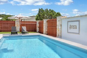 Una piscina con dos sillas de descanso y una sombrilla, rodeada por una cerca de madera y un muro de piedra con la palabra "ARTISTA".