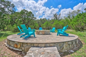 A stone fire pit with four blue Adirondack chairs arranged around it, set in a circular stone platform surrounded by trees.
