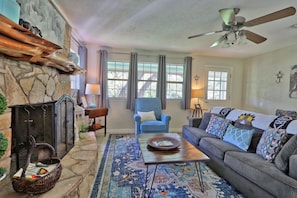 A cozy living room with a stone fireplace, a blue armchair, a sofa with patterned cushions, and a large rug.