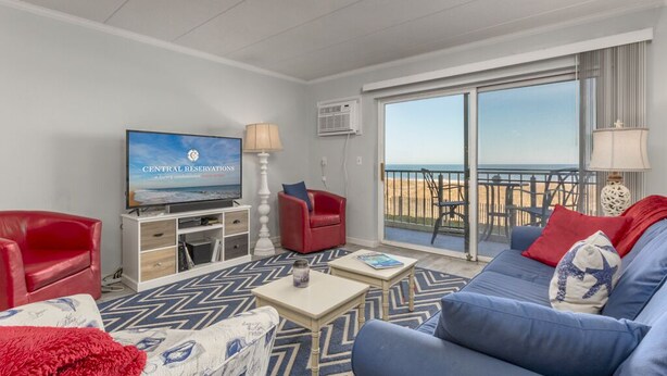 A modern living room with a television, red and blue sofas, a patterned rug, and a balcony with a view. - Well maintained 1 BR, 1.5 BA oceanfront condo. Enjoy the beautiful ocean views from your balcony and inside the unit. (Ocean City)