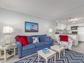 Un salon avec un canapé bleu, une table basse et un fauteuil à motifs. Au fond, on aperçoit une cuisine avec des meubles blancs et un four à micro-ondes.