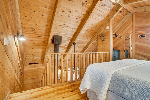 Cozy cabin-style room with a high ceiling and exposed beams, featuring a bed with a quilted cover, a wooden cabinet, and a hanging light fixture.