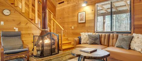 A cozy cabin interior with a fireplace, a sofa, a chair, and a staircase.
