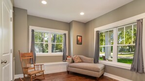 A room with two large windows, a rocking chair, a sofa, and a picture on the wall. - Four Generations (Saugatuck)