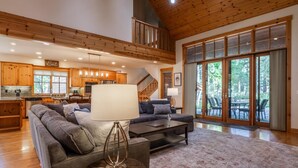 Ein geräumiger Wohnbereich mit einer großen Couch, einem Couchtisch und einem Esstisch. Im Hintergrund befindet sich eine Küche mit Holzschränken und eine Treppe, die zu einer oberen Ebene führt.