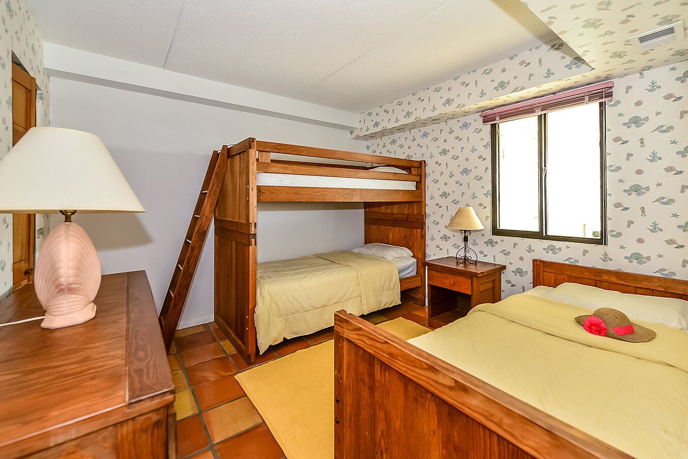 A room with a bunk bed, a desk with a lamp, a window with curtains, and patterned wallpaper.