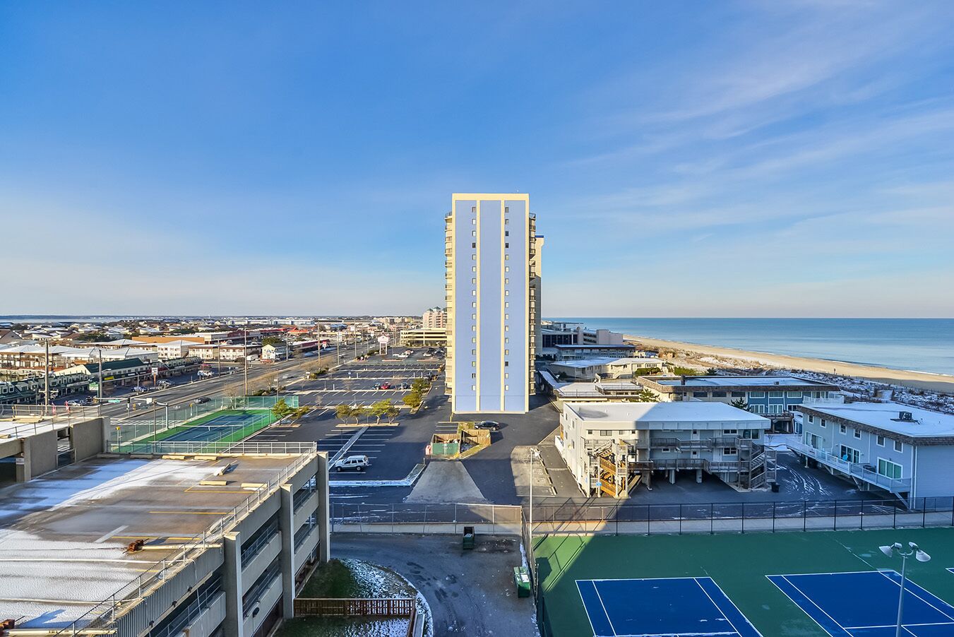 Ein Hochhaus mit mehreren Stockwerken, umgeben von einem Parkplatz und Tennisplätzen, mit Blick auf einen Strand.