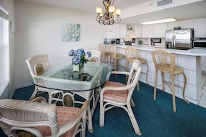 A dining area with a glass table surrounded by wicker chairs, a kitchen with white cabinets and stainless steel appliances, and a blue carpet.