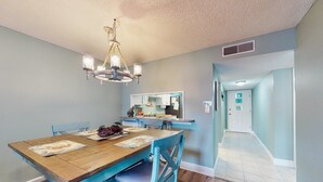 Un coin repas avec une table en bois et des chaises bleues, un lustre et une cuisine en arrière-plan.
