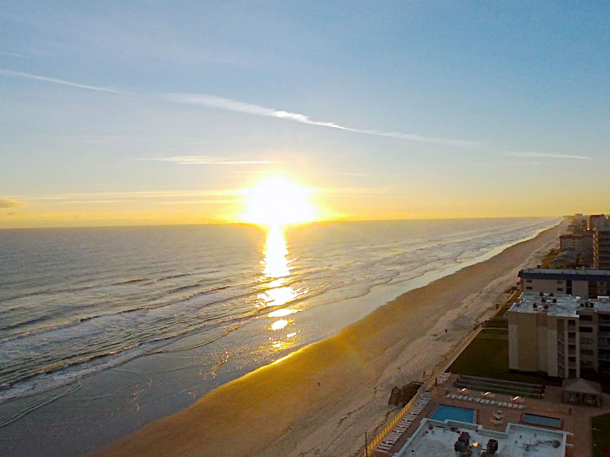 Blick auf einen Strand bei Sonnenuntergang, ein Poolbereich und angrenzende Gebäude.