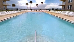 Une piscine entourée d’une clôture et de bâtiments, avec des chaises longues et des palmiers.
