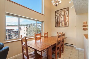 A dining area with a wooden table and chairs, large windows with a view of buildings and trees, and a chandelier. - Beautiful Breckenridge views, Close to lifts/downtown, Garage parking! (Breckenridge)