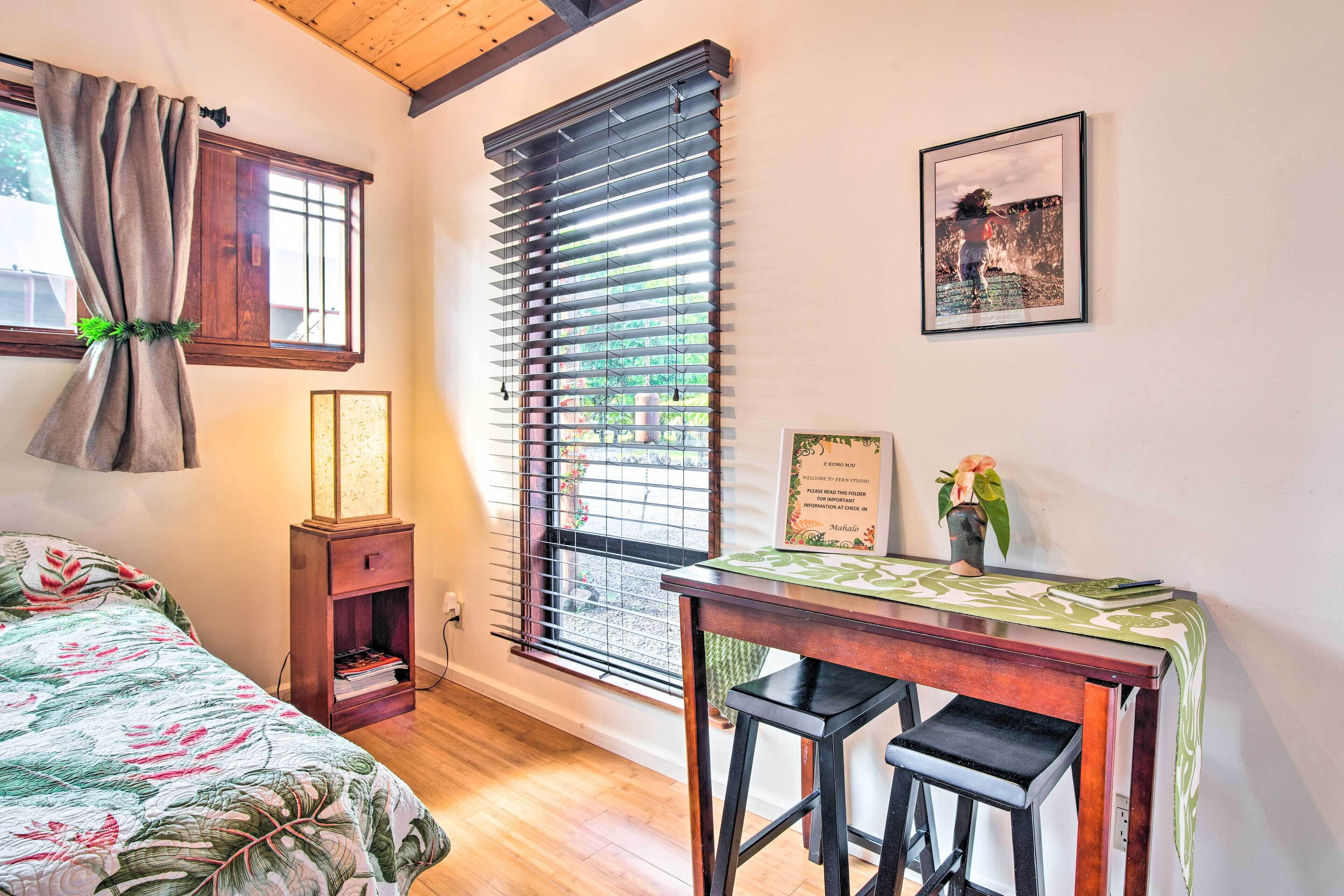 Une pièce comprenant un lit, un bureau avec deux chaises, une fenêtre avec des stores, un tableau encadré au mur et une petite table sur laquelle se trouvent un vase et un livre.