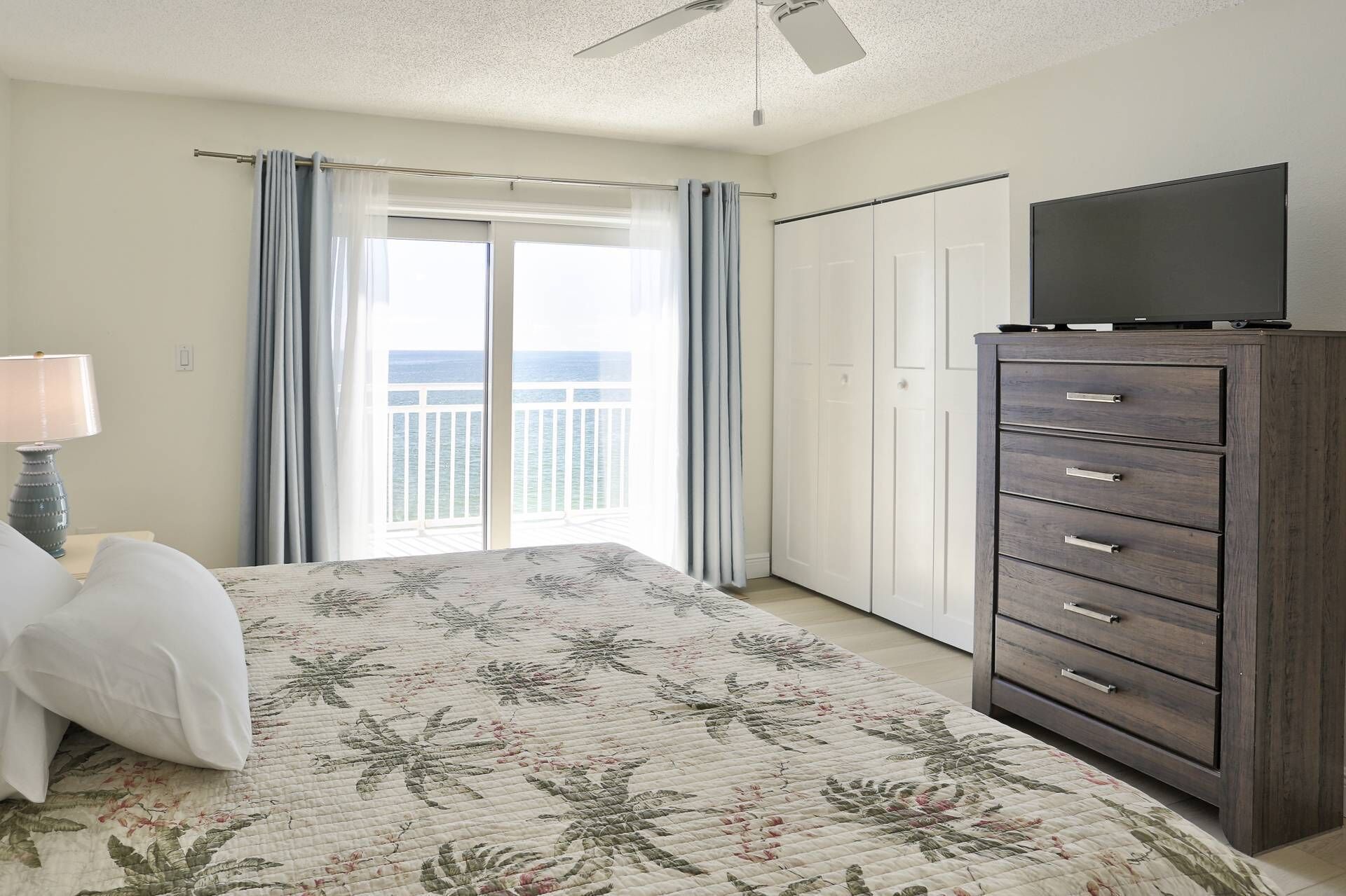 A bedroom with a bed, a dresser, a television on top, and a view of the ocean through a sliding door.
