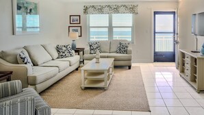 A living room with a beige sofa, a coffee table, and a patterned rug. There are two windows with blinds, a door, and a television on a stand.
