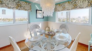 A dining area with a round glass table surrounded by white wicker chairs, a chandelier, and a view of the city through large windows.