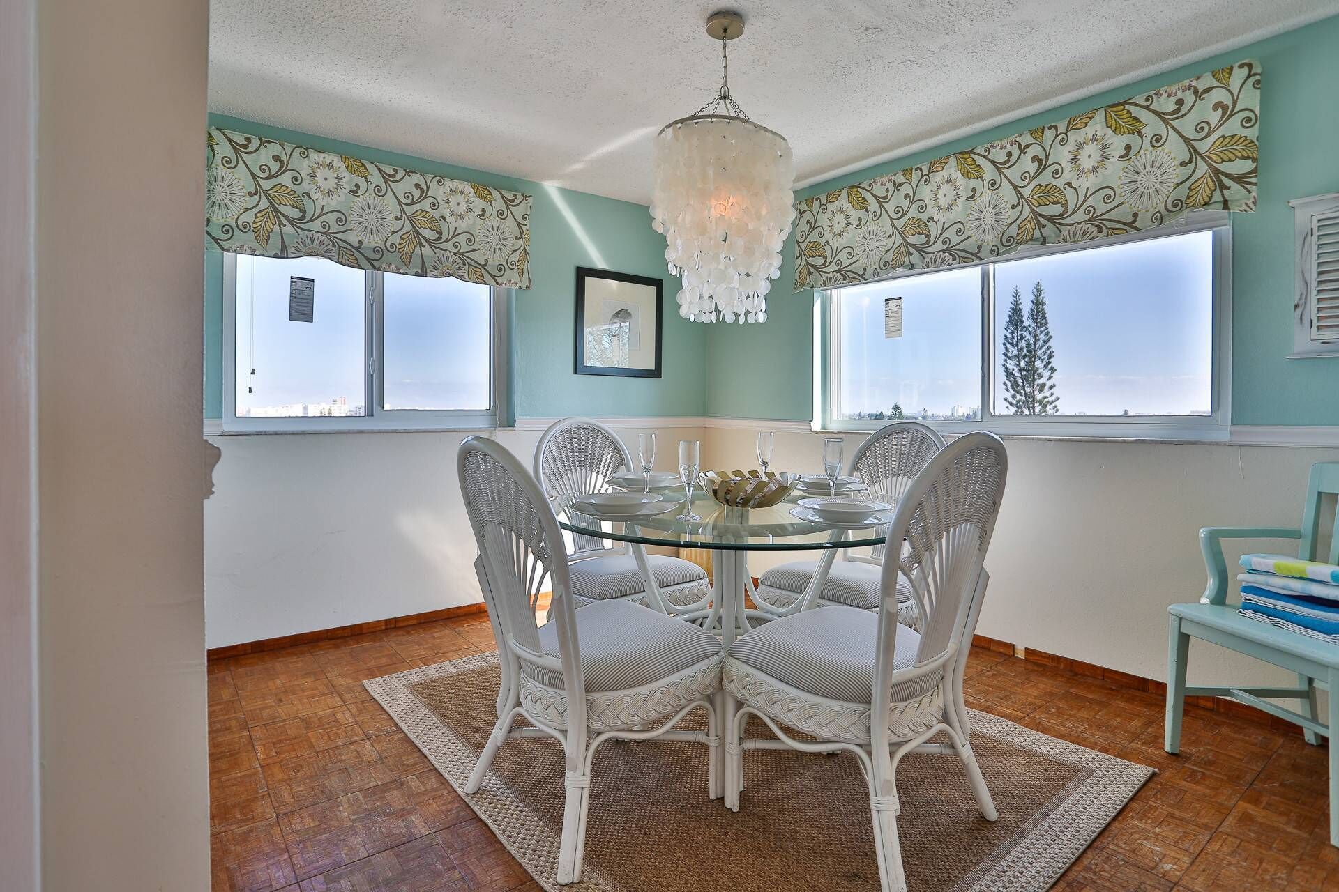 A dining room with a round glass table, white wicker chairs, a chandelier, and patterned curtains.
