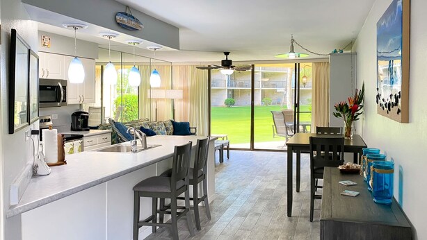 A modern kitchen with a central island, a dining area with a table and chairs, and a view of a green lawn through large windows.