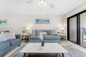 Una sala de estar con un sofá gris, una mesita de centro blanca y un cuadro que representa una playa.
