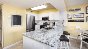 A kitchen with white cabinets, a stainless steel refrigerator, a microwave, and a granite countertop. - PI 406: Sunset Shores - Take A Look! Perfect Beach Retreat with Beach Service (Fort Walton Beach)