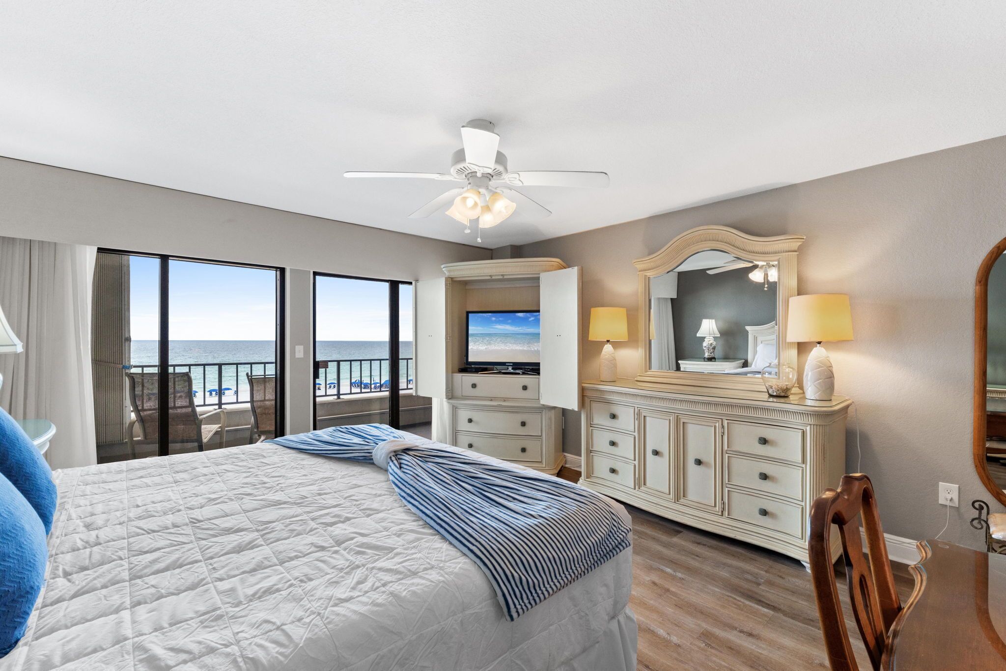 Habitación de hotel con cama, televisor, cómoda, espejo y balcón con vista al océano.