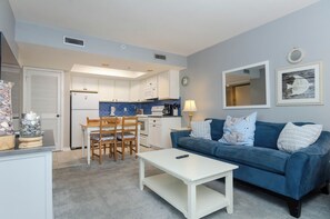 A compact living space with a kitchenette, a dining area with a table and chairs, a blue sofa with decorative pillows, and a small white coffee table.