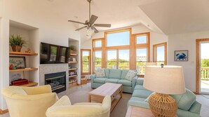 A spacious living room with a fireplace, a ceiling fan, a large TV, and a view of the outdoors. - Rookery - Corolla, Whalehead, 395 Yds to Beach, Ocean View, Pool, Hot Tub, Pets OK, Flex Stays (Corolla)