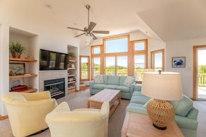 A spacious living room with a fireplace, a ceiling fan, a large TV, and a view of the outdoors. - Rookery - Corolla, Whalehead, 395 Yds to Beach, Ocean View, Pool, Hot Tub, Pets OK, Flex Stays (Corolla)