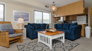 Un espace de vie compact comprenant un canapé bleu, une table basse en bois, un panier en osier, une chaise à rayures et un coin cuisine avec des meubles en bois et un réfrigérateur blanc.