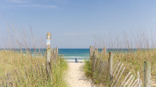 Un sentier qui longe la plage et mène à l’océan, avec une clôture en bois et un panneau indicateur.