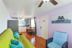 A room with a green sofa, a wicker chair, a dining table with chairs, a kitchen area, and a door with a maritime painting. - Steps to Private Beach! Pet-Friendly Getaway (Galveston)