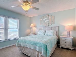 A bedroom with a bed, two bedside tables, a ceiling fan, a window with blinds, and wall art. - Perfect view of the Ocean! Directly on No Drive Beach of New Smyrna (New Smyrna Beach)