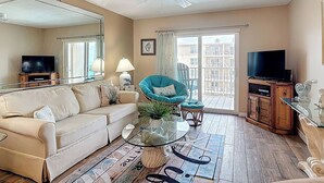 Ein Wohnzimmer mit einer Couch, einem Glastisch, einem blauen Sessel, einem Fernseher und einem Balkon mit Blick auf Gebäude.
