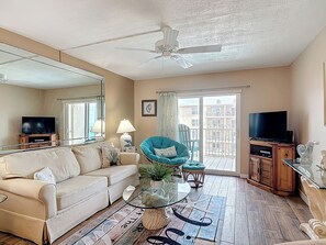 A living room with a sofa, a glass coffee table, a blue armchair, a television, and a balcony with a view of buildings. - Perfect view of the Ocean! Directly on No Drive Beach of New Smyrna (New Smyrna Beach)