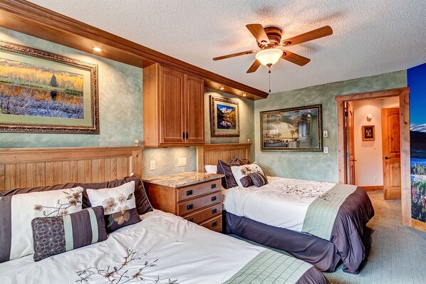 A bedroom with two beds, wooden headboards, a ceiling fan, and artwork on the walls.